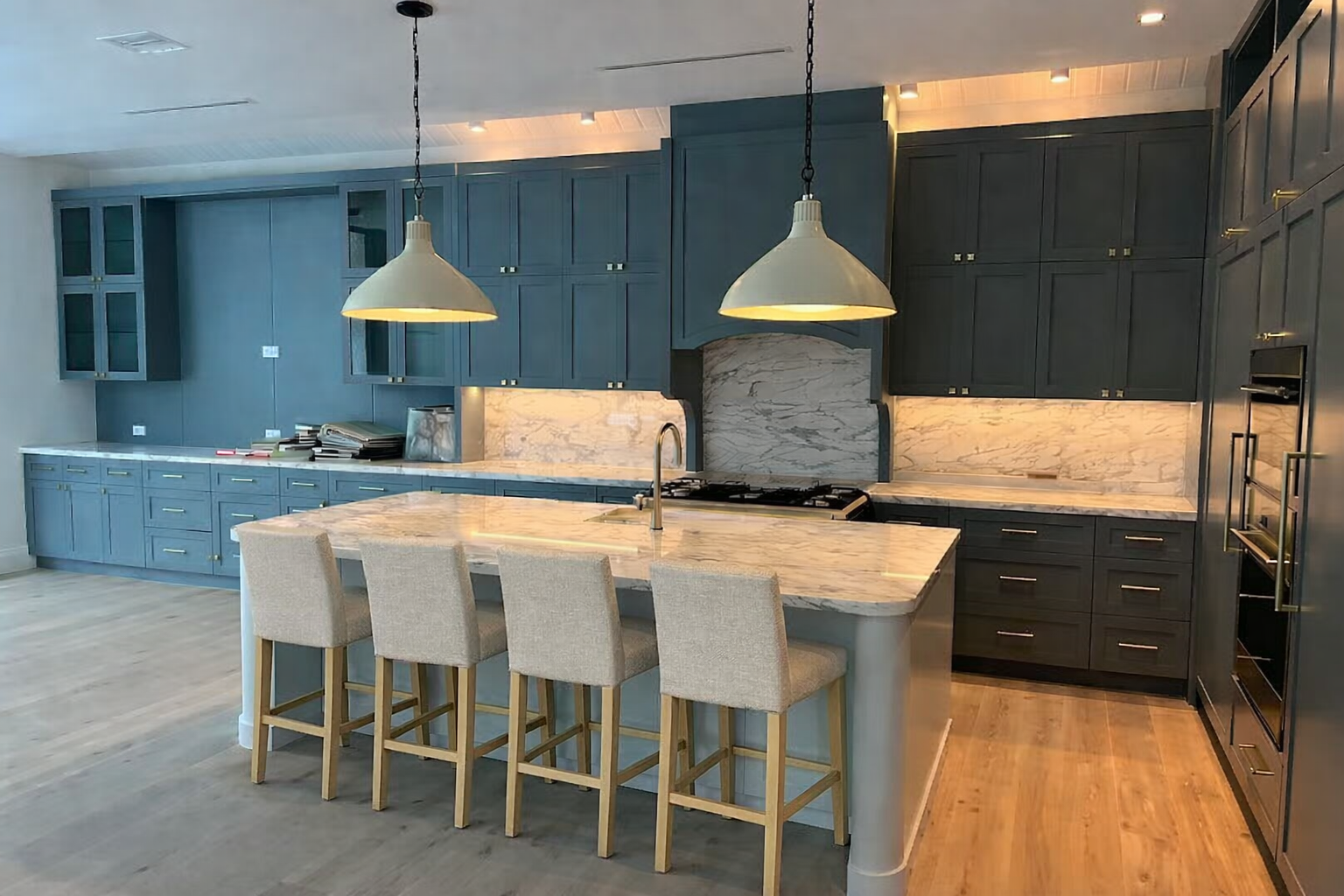 Modern kitchen with dark blue cabinets, marble countertops, and a marble backsplash. There are four beige bar stools at the island, with two large pendant lights hanging above.