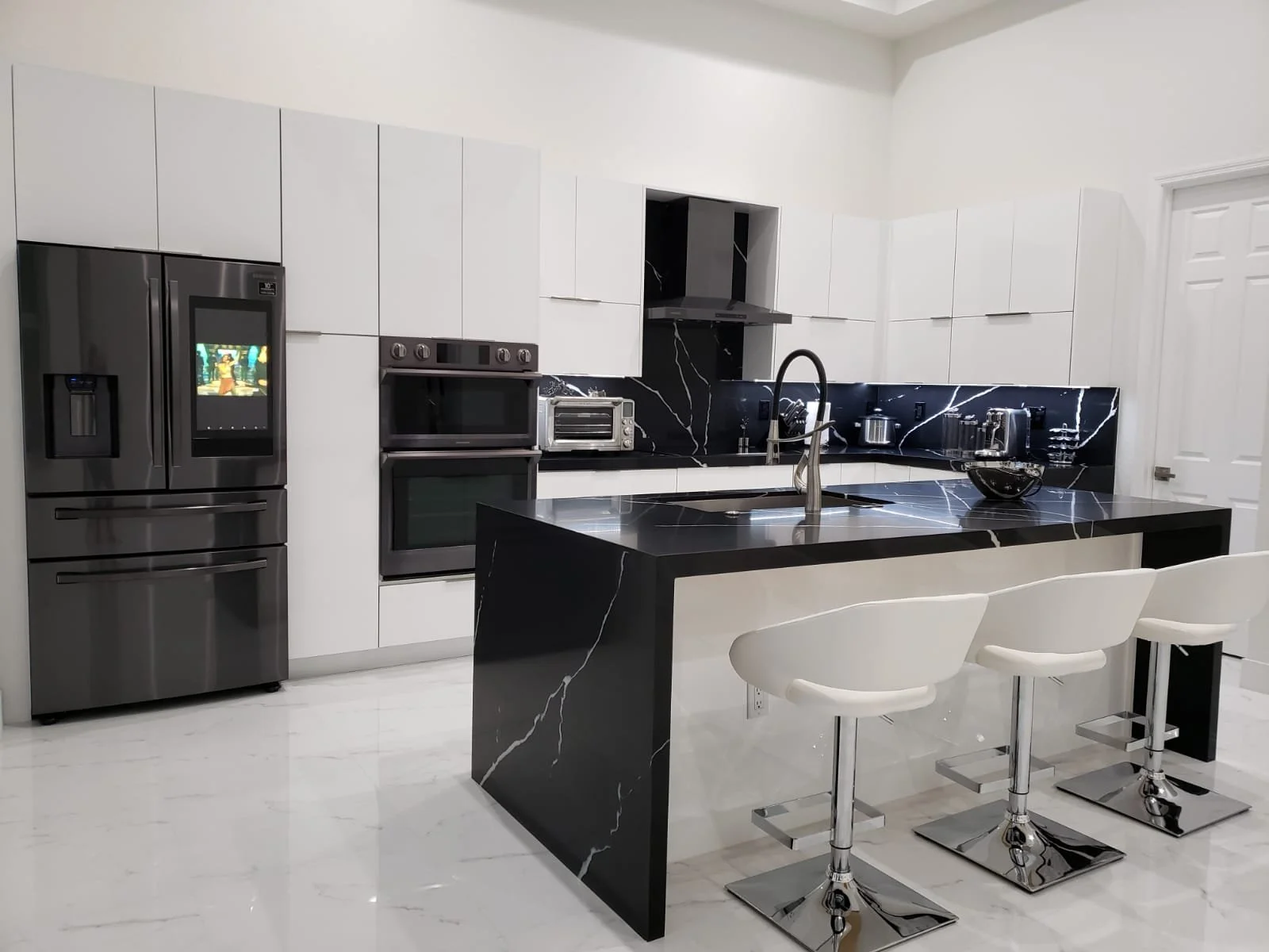 Modern kitchen with white cabinets, black marble countertops and island, stainless steel appliances, and three white barstools.
