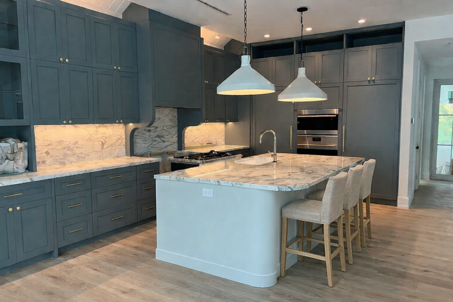 Modern kitchen with blue cabinets, marble countertops, and a central island with seating. There are two hanging pendant lights above the island, a built-in oven, and a gas stove with a marble backslash. Wood flooring and a door leading outside are visible.