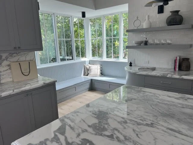 Bright kitchen with gray cabinets, marble countertops, and a bay window corner seating area with a cushion and view of green trees outside.
