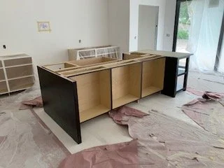 Unfinished kitchen island with open shelves, surrounded by wrapping paper and debris, in a room with white walls and large windows.
