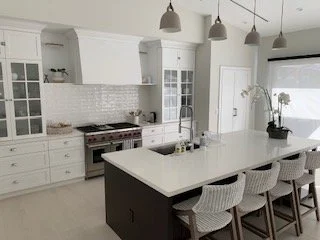 Modern kitchen with white cabinetry, an island with a dark base, and four textured barstools. White backsplash, stainless steel appliances, and pendant lights above the island.