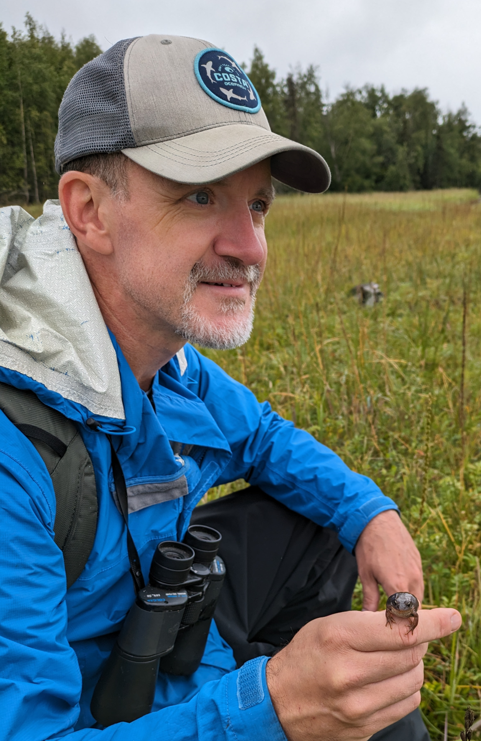 A man outdoors in a grassy field, wearing a blue jacket, gray cap with a 'COSTA' logo, and binoculars around his neck, holding a small amphibian or reptile in his hand.