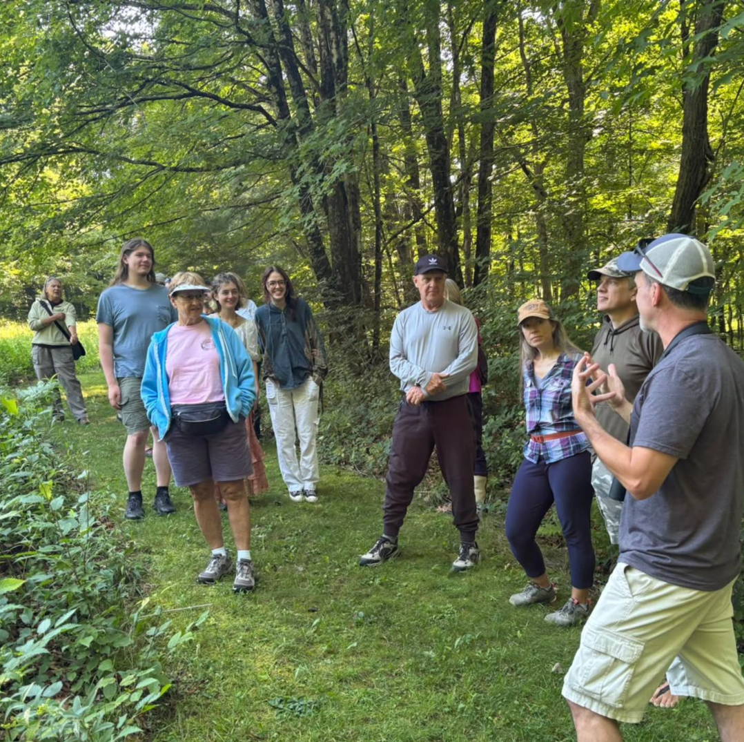 Group of people listening to a guide during a nature walk in the woods, surrounded by trees and greenery.