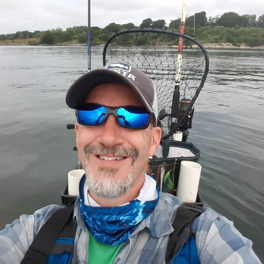 Man in a kayak taking a selfie with a smile, wearing sunglasses, a cap, a blue bandana, and a fishing rod in the water behind him, with trees and cloudy sky in the background, on the ocean