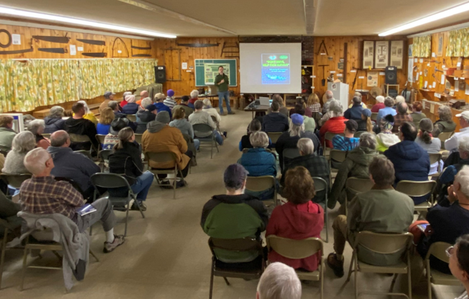 A large group of people sitting in chairs, attending a presentation in a room with wooden walls and various tools and items hanging on the walls. A man stands in front of a screen giving a talk.