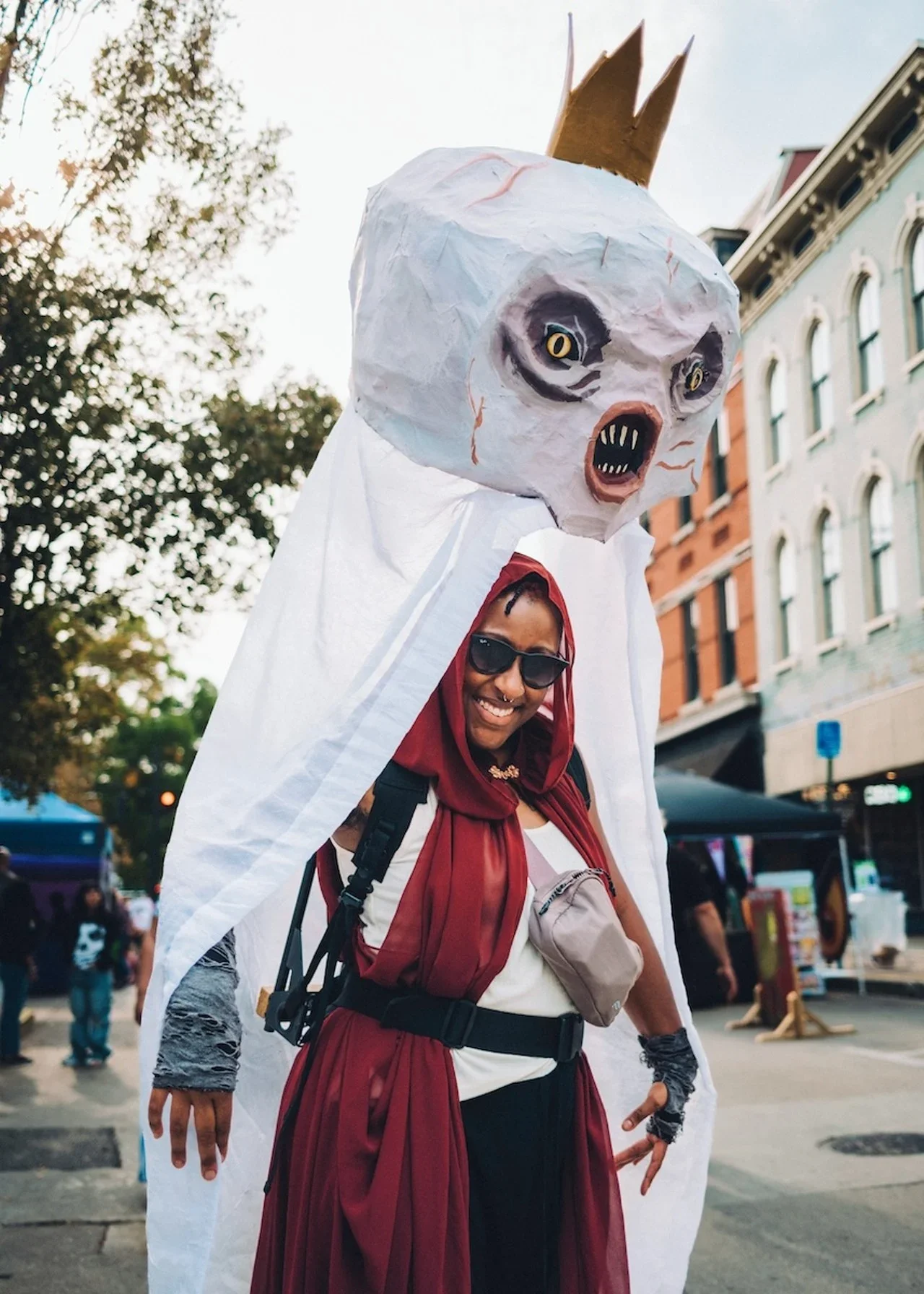 a black woman wears a giant body puppet of a white, fanged cryptid