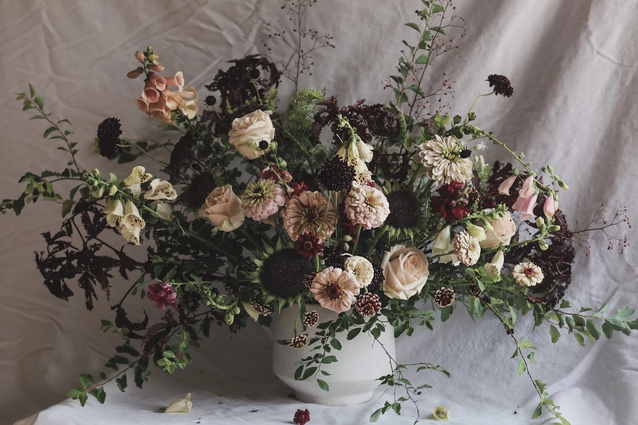 A large bouquet of various flowers in a white ceramic vase, placed on a draped white cloth background.