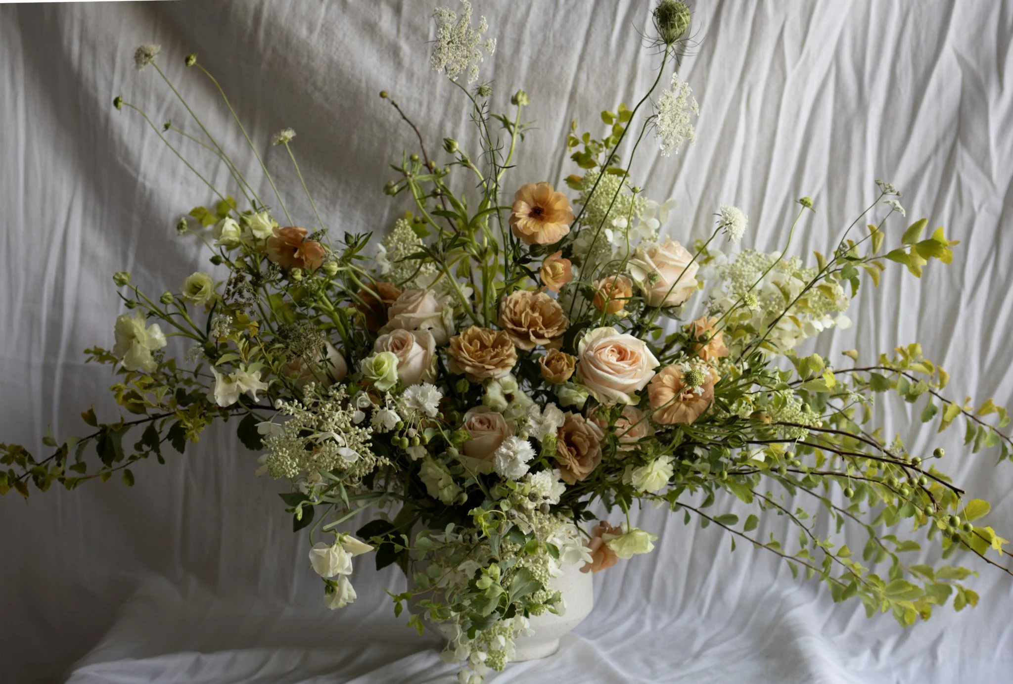 A large floral arrangement with peach, white, and cream-colored roses, white lisianthus, and various green foliage, set against a crinkled white fabric background.