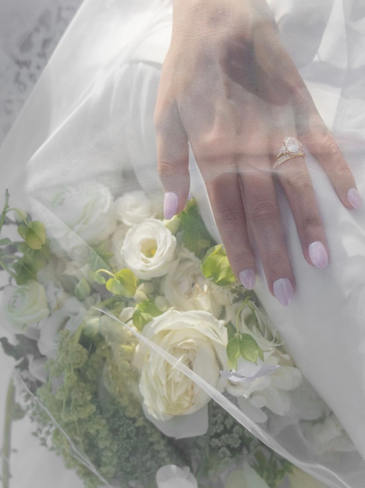 A woman's hand with pink and white nails, wearing a ring with a large gemstone, resting on a bouquet of white roses and green foliage, partially covered by a sheer white fabric.