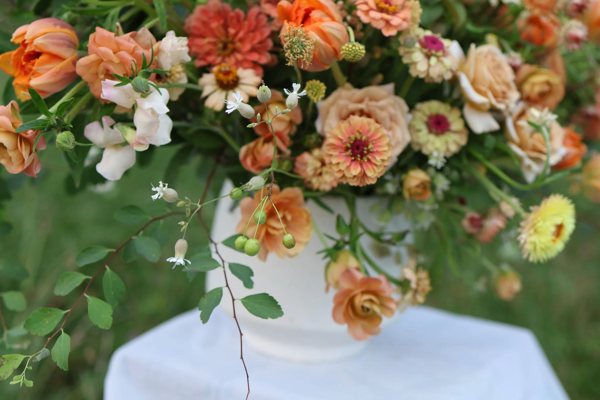 A bouquet of various flowers including orange and pink roses, small white flowers, and orange zinnias, arranged in a white vase with a blurred green background.