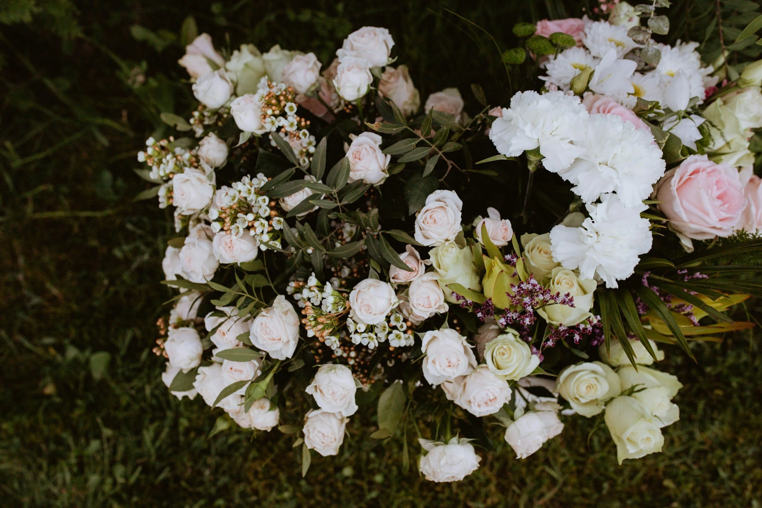 A floral arrangement with white, pink, and yellow roses, white carnations, small white and purple flowers, and green foliage on grass.