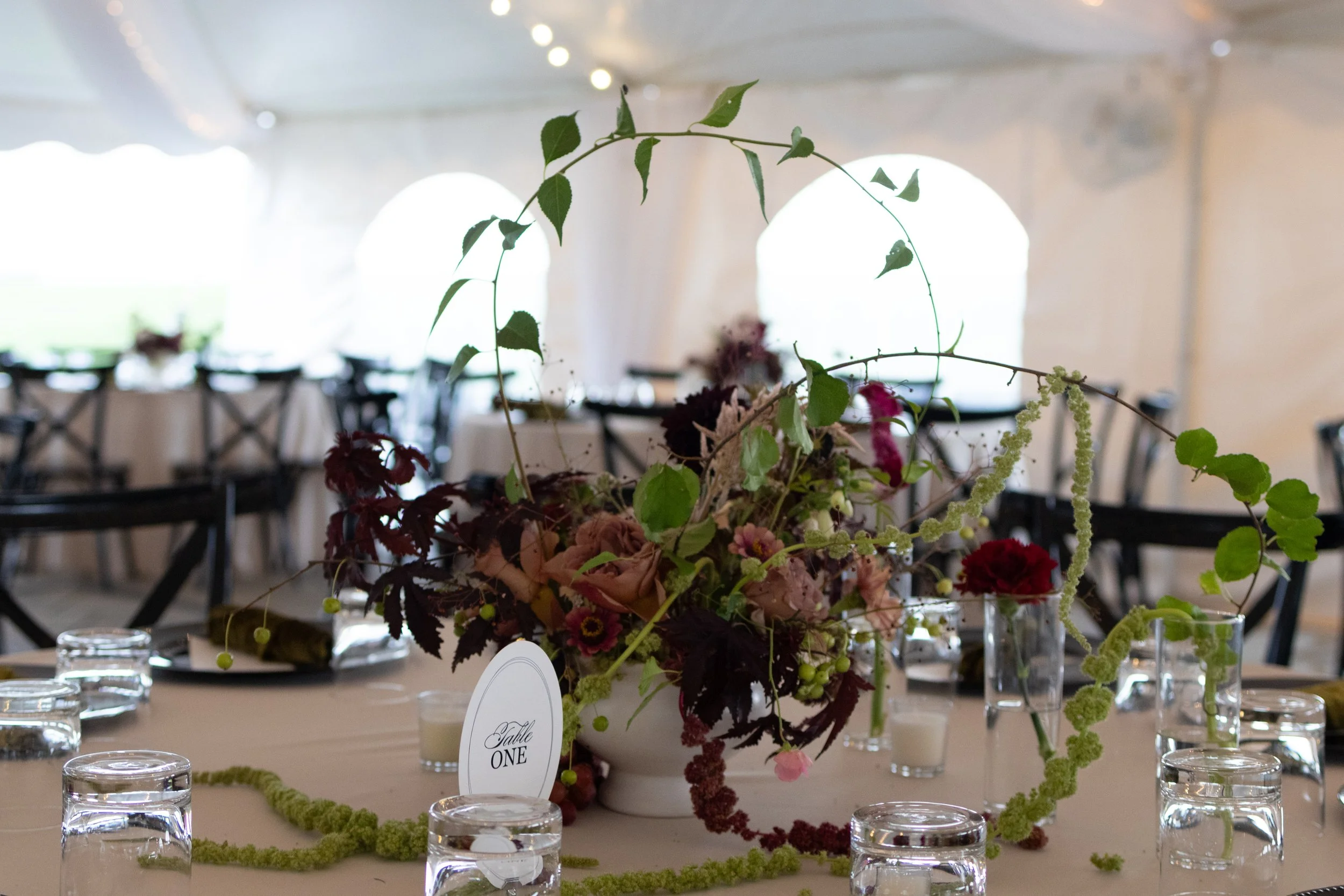 Elegant banquet table featuring a floral centerpiece with green, pink, and purple flowers, surrounded by water glasses, set in a warmly lit room with arched windows.