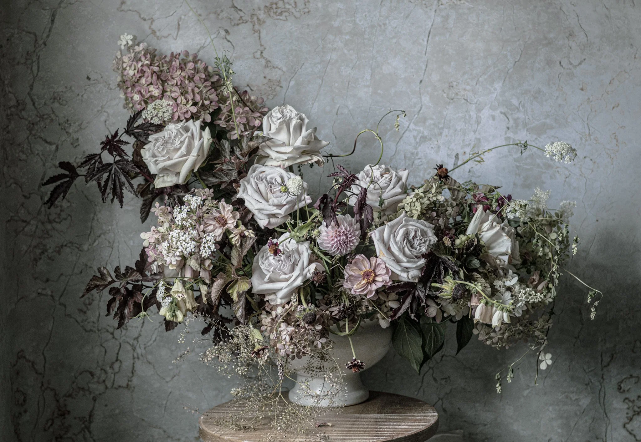 A large bouquet of pastel-colored flowers, including roses and hydrangeas, placed in a white vase on a wooden stand against a textured gray wall.