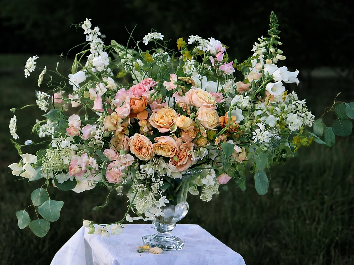 A large, colorful flower arrangement in a clear glass vase on a white cloth-covered table outdoors. The arrangement includes peach roses, pink carnations, white orchids, small white flowers, and green foliage.