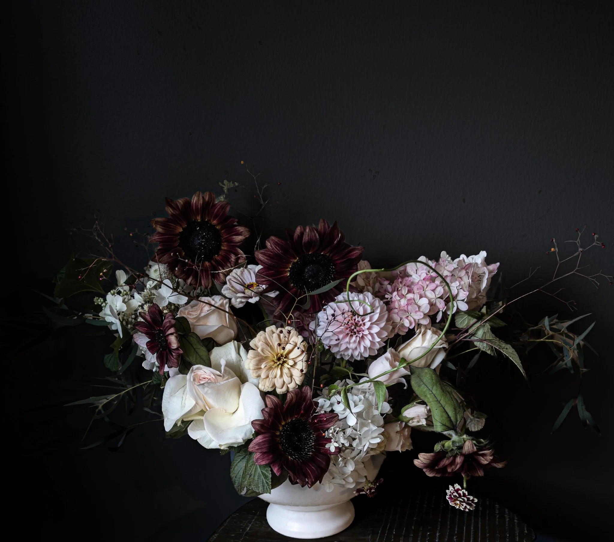 A bouquet of dark red, white, and pink flowers in a white vase against a dark background.