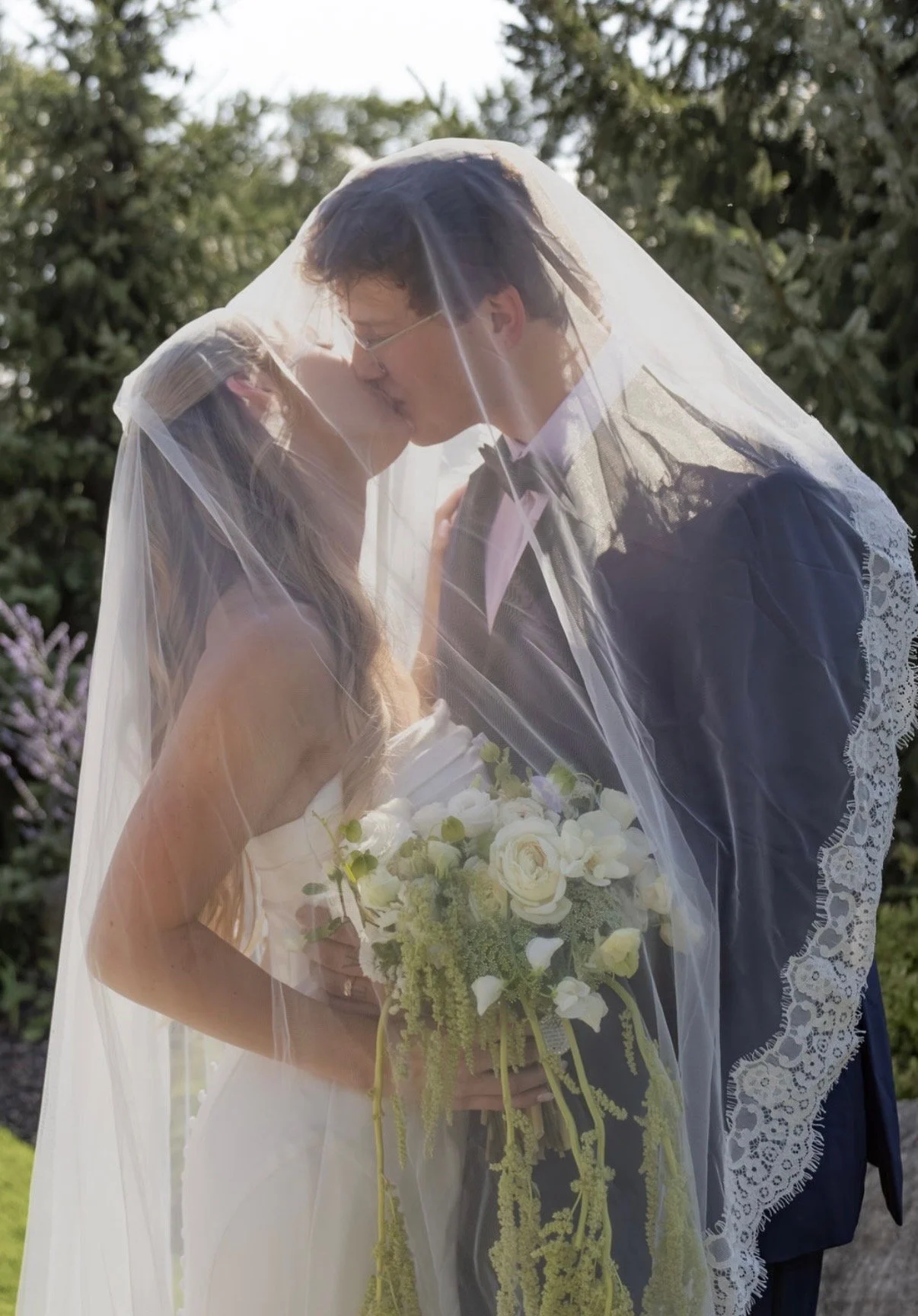 A bride and groom kissing under a lace veil during their wedding, with the bride holding a bouquet of white flowers and greenery outdoors.