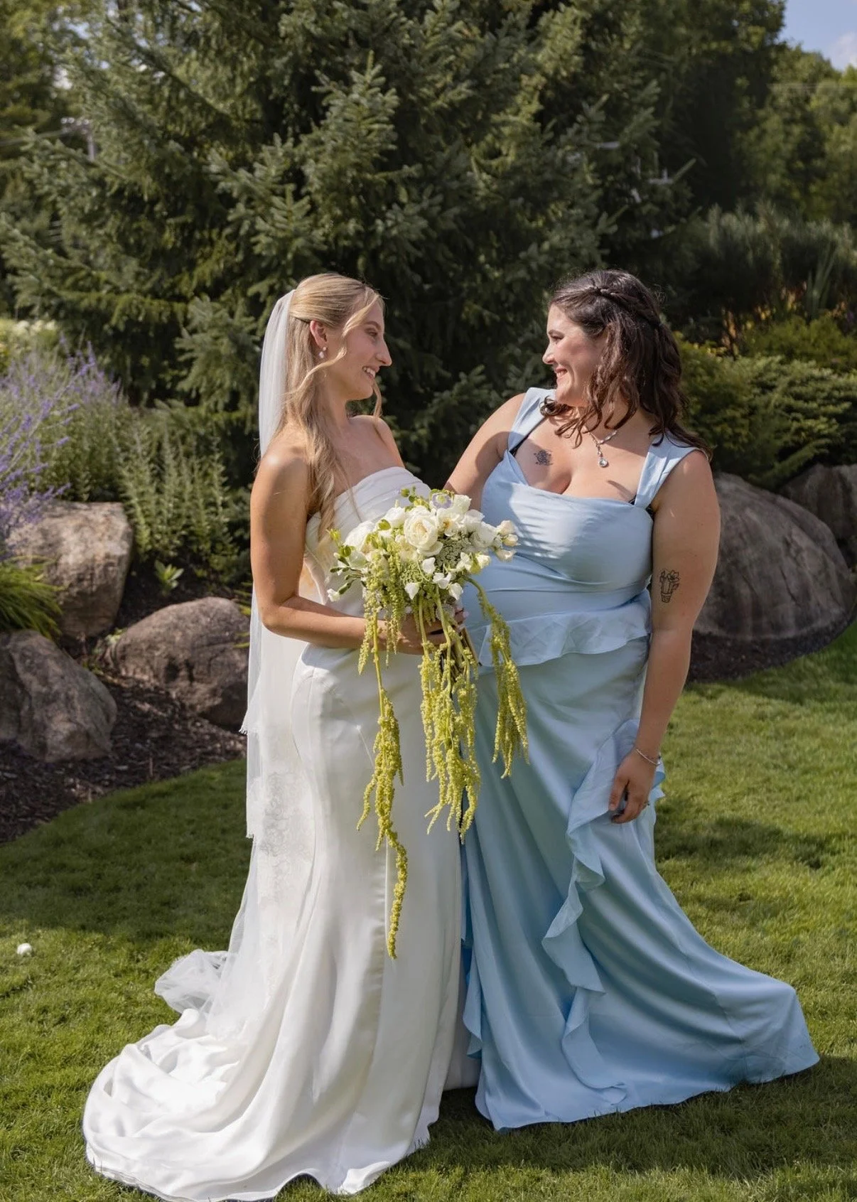 Two women, one in a white wedding dress holding a bouquet, and the other in a light blue dress, smiling at each other outdoors in a lush garden with trees and rocks.