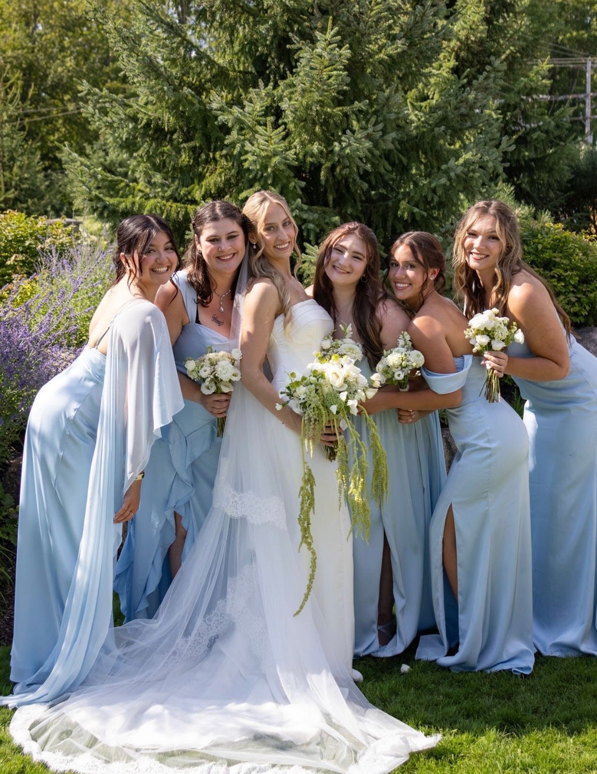A bride and six bridesmaids in light blue dresses, holding bouquets of white flowers, standing outdoors with lush green trees in the background.