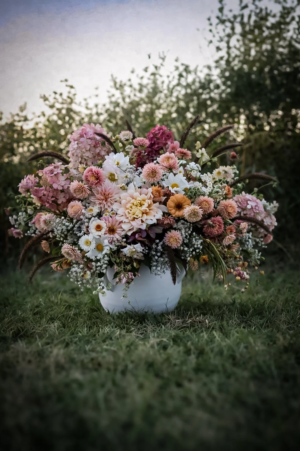 A white vase filled with a large assortment of pink, white, and peach flowers is placed on the grass outdoors, with a blurred background of trees and sky.