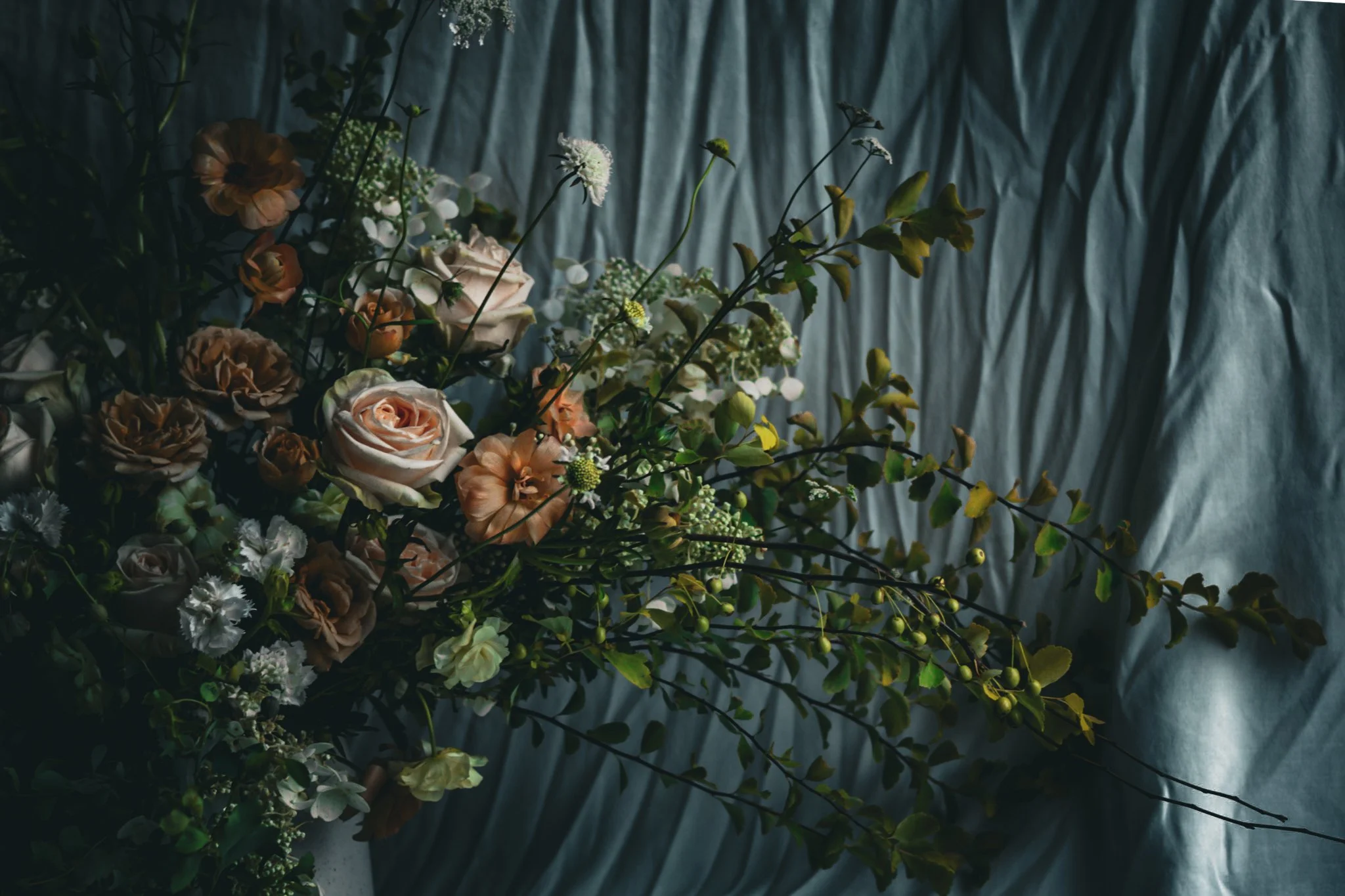 A close-up of a flower arrangement with roses, white flowers, and green foliage set against a dark textured background.