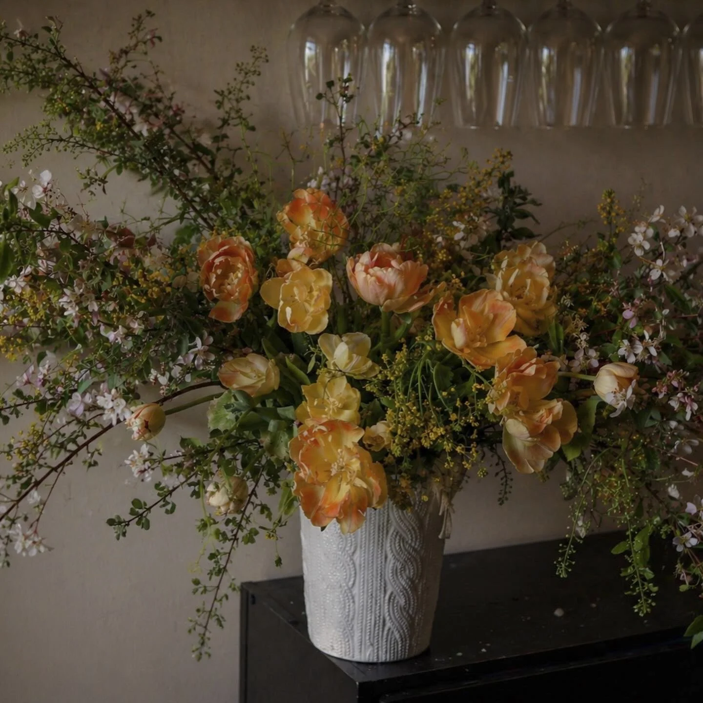 A large floral arrangement of yellow and orange tulips, pink and white small flowers, and greenery in a tall white textured vase on a black surface.