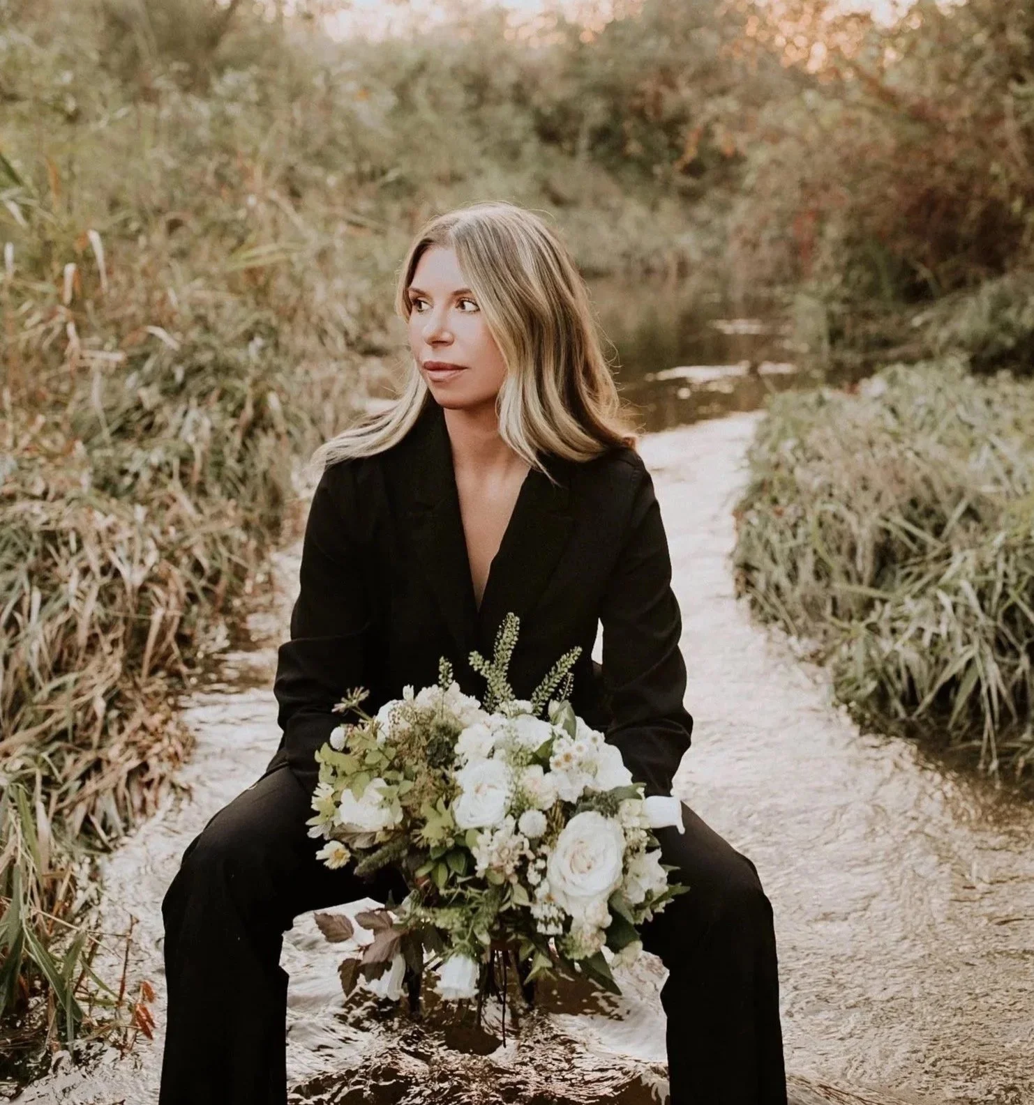 A woman sitting on a narrow dirt path next to a stream, holding a bouquet of white flowers, with autumn foliage in the background.
