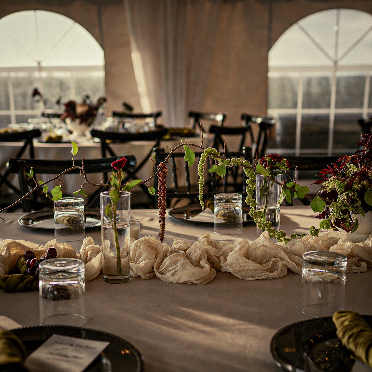 Elegant table setting with vases of greenery, candles, and decorative fabric, set in a tent with large windows