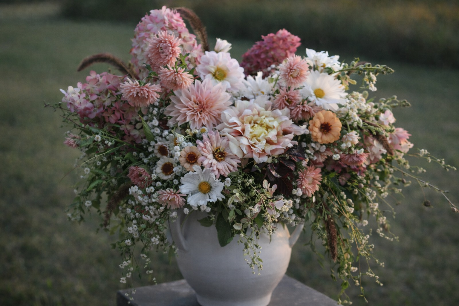 A large bouquet of pink, white, and peach flowers in a white ceramic vase on a stone surface outdoors.