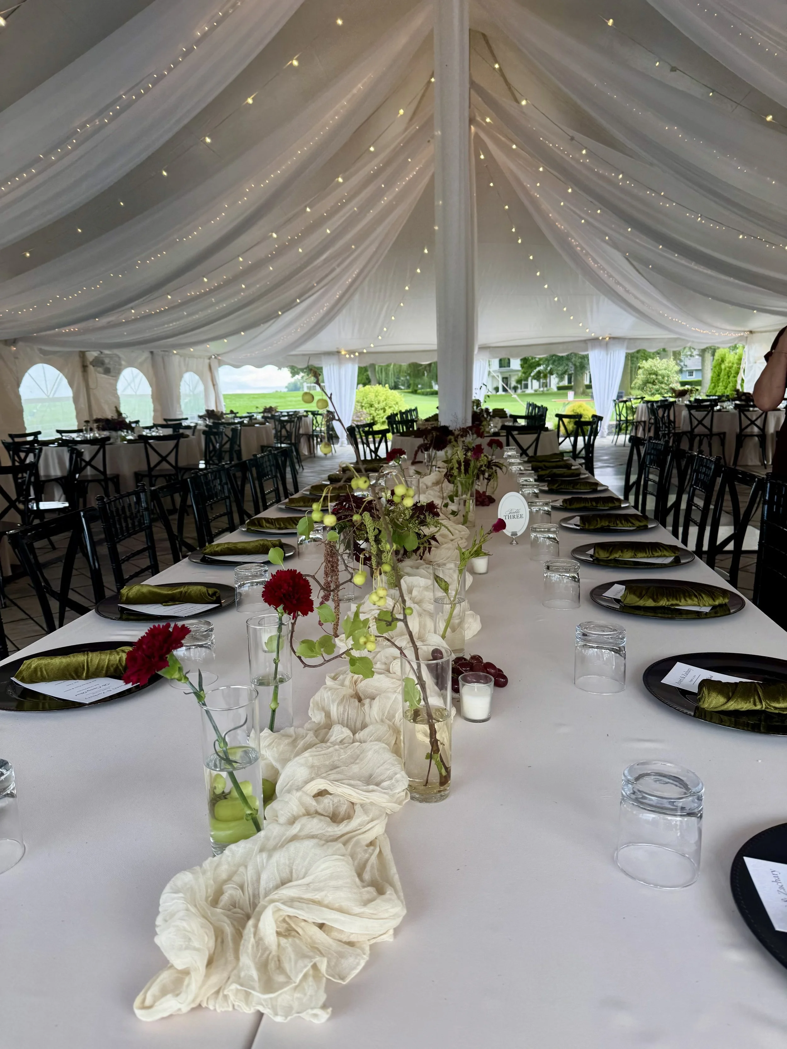 A decorated outdoor event tent with a long banquet table set with black plates, green napkins, clear glasses, and floral centerpieces with red and green flowers, candles, and a beige fabric runner. The tent has draped white fabric and string lights, 