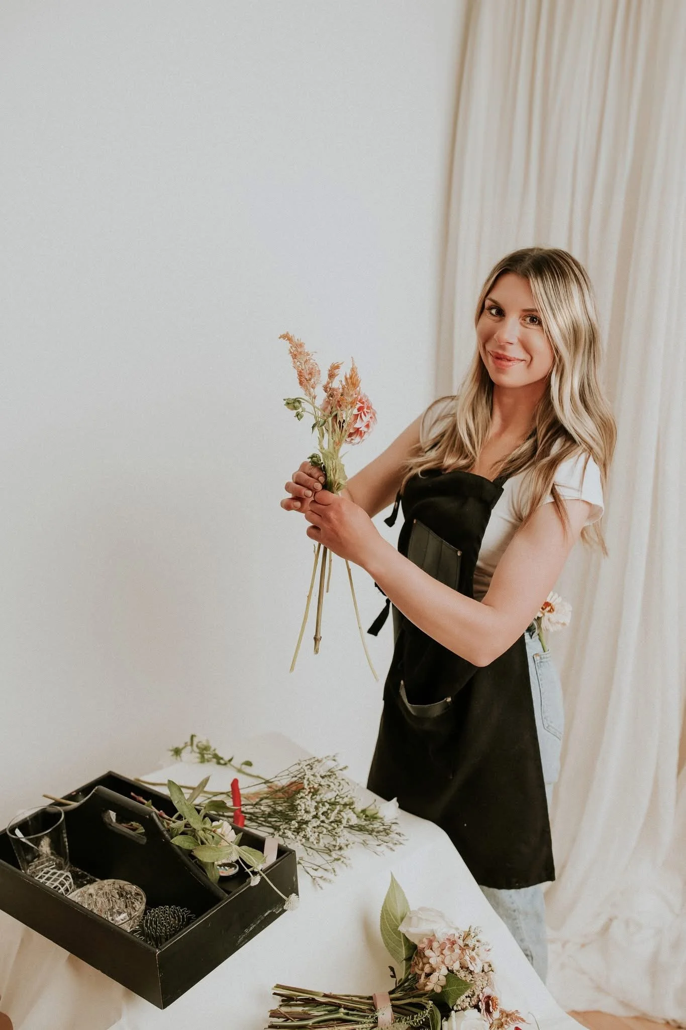 A woman with wavy blonde hair and a black apron is arranging flowers at a floral workshop. She is holding a bouquet of pink and beige flowers, with greenery. The workspace has flowers, floral supplies, and tools on a white table.