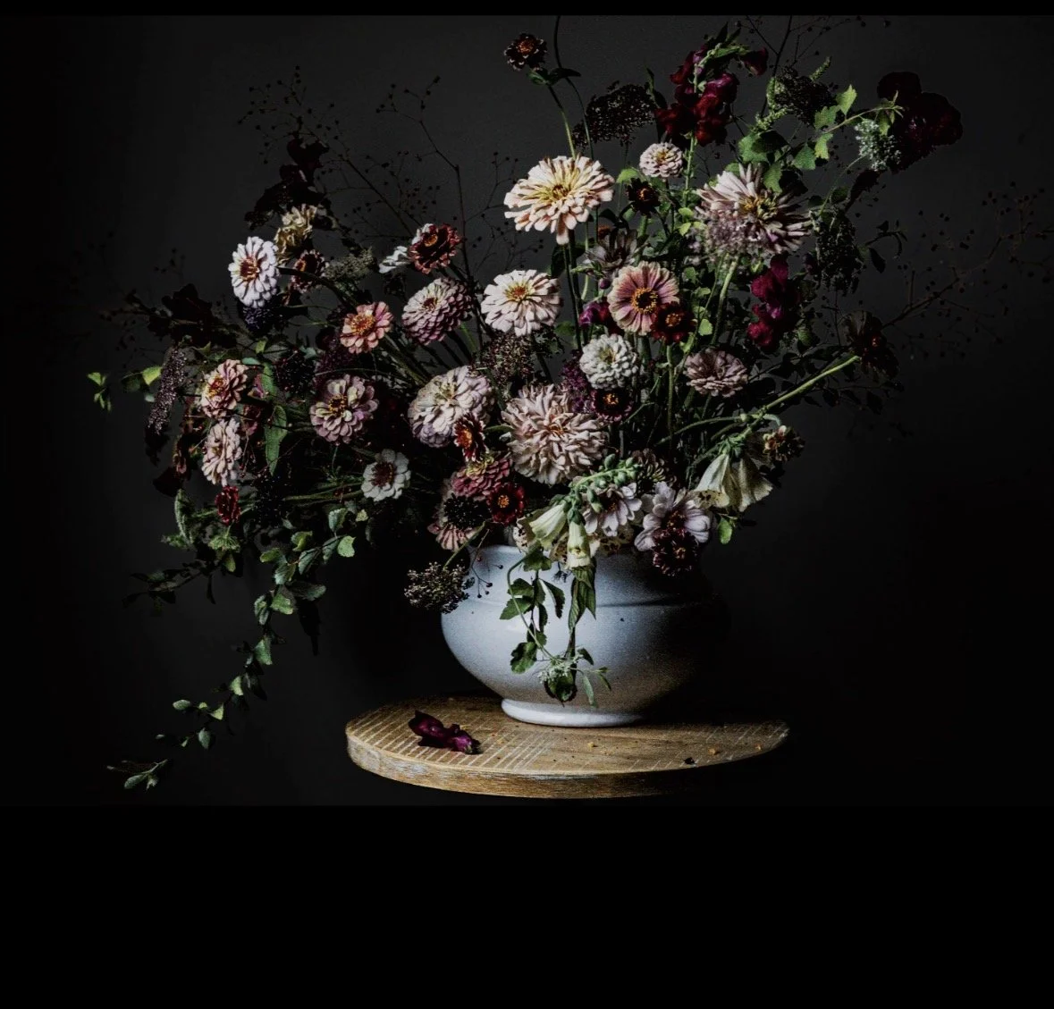 A white vase with a large bouquet of assorted flowers, including pink, white, and dark red blooms, placed on a round wooden table against a dark background.