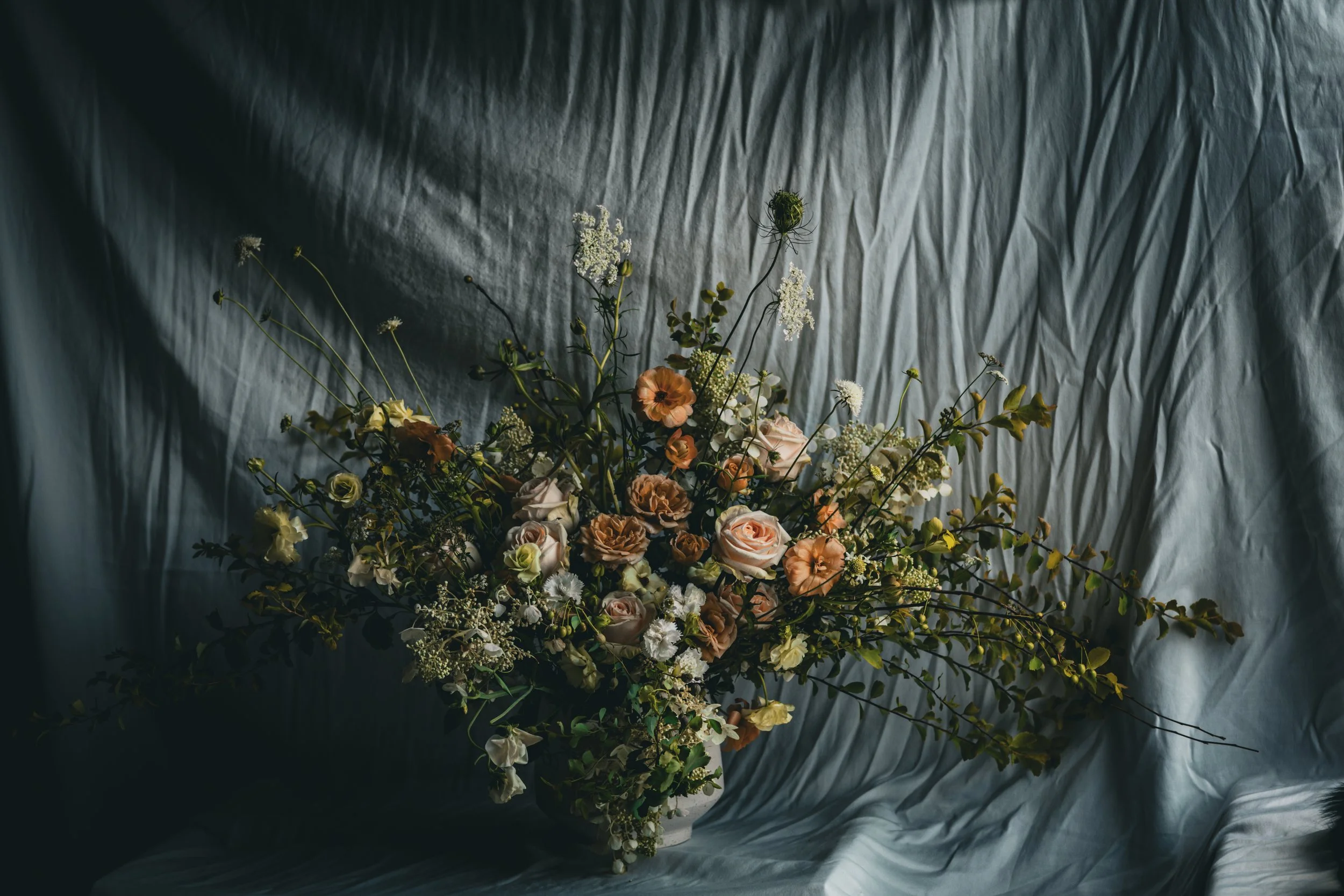 A floral arrangement with various flowers including roses, carnations, and other small white flowers, set against a textured, dark background.