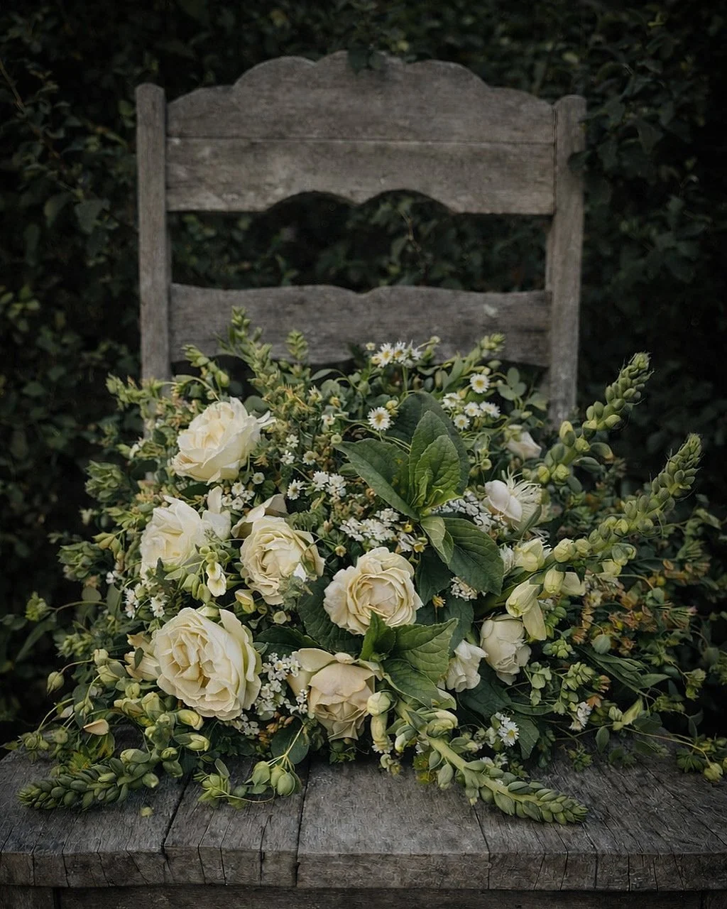 A bouquet of white roses, small white daisies, and greenery placed on a rustic wooden table in front of an old wooden chair outdoors.