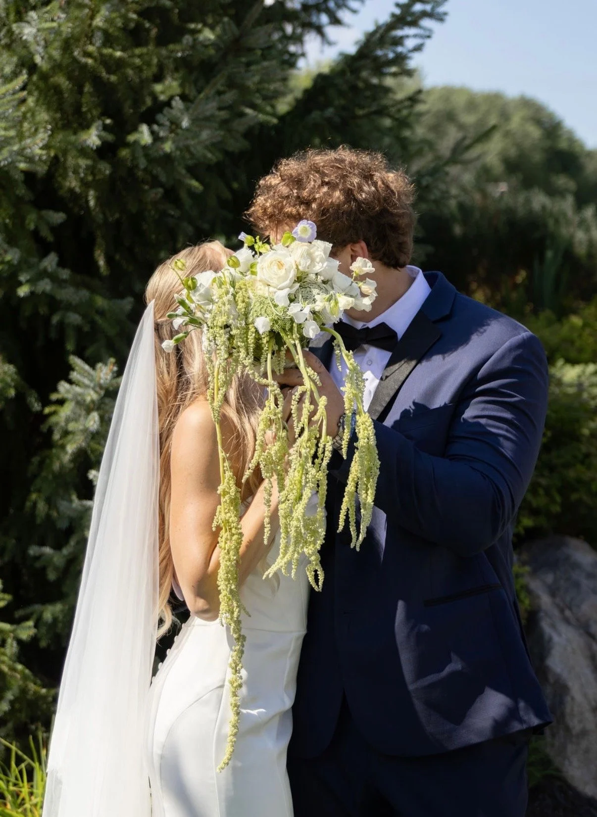 A bride and groom sharing a kiss during their wedding ceremony outdoors, with the groom holding a large bouquet of white flowers with green accents, in front of a lush greenery background.