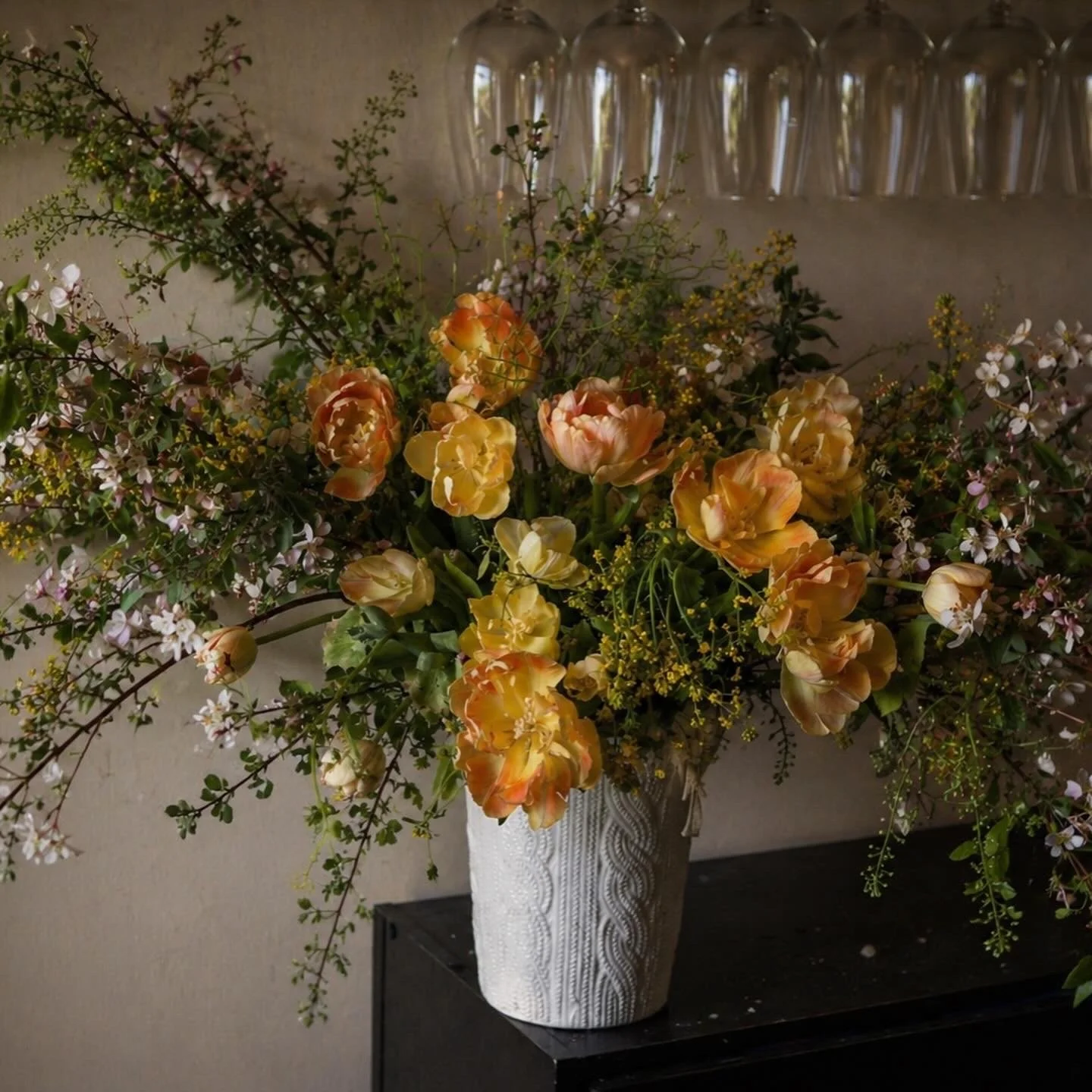 A large floral arrangement with yellow and orange tulips, white flowers, and green foliage in a tall, white textured vase on a black table.