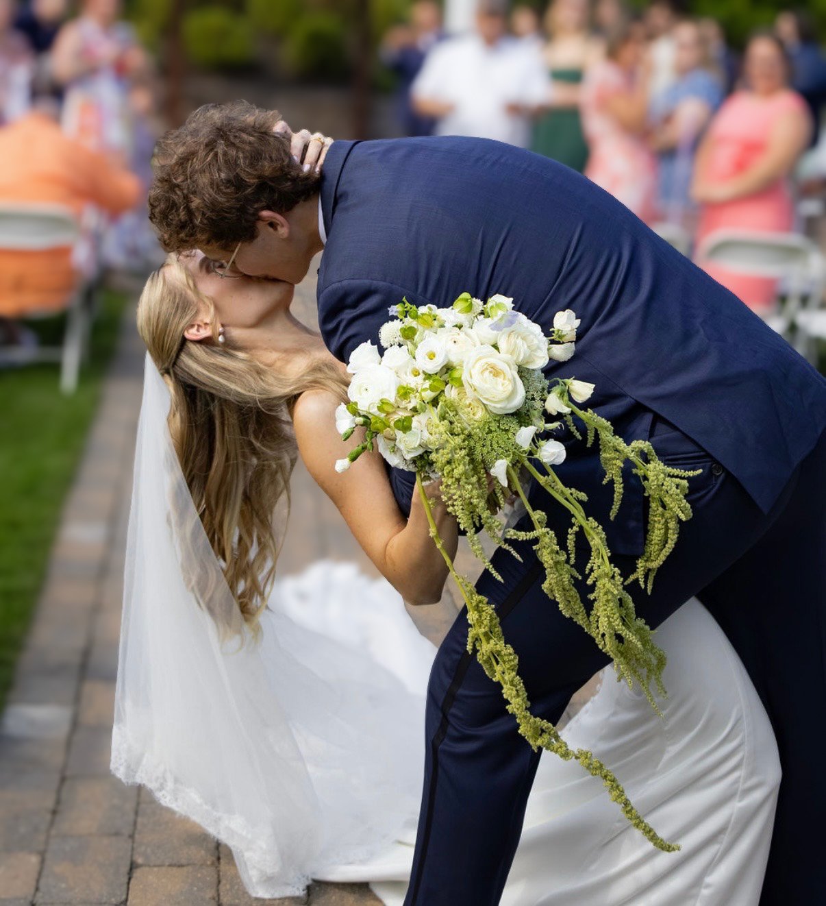 A bride and groom share a kiss at their wedding, with the groom bending down to kiss the bride, who is holding a large bouquet of white flowers. Guests are visible in the background.