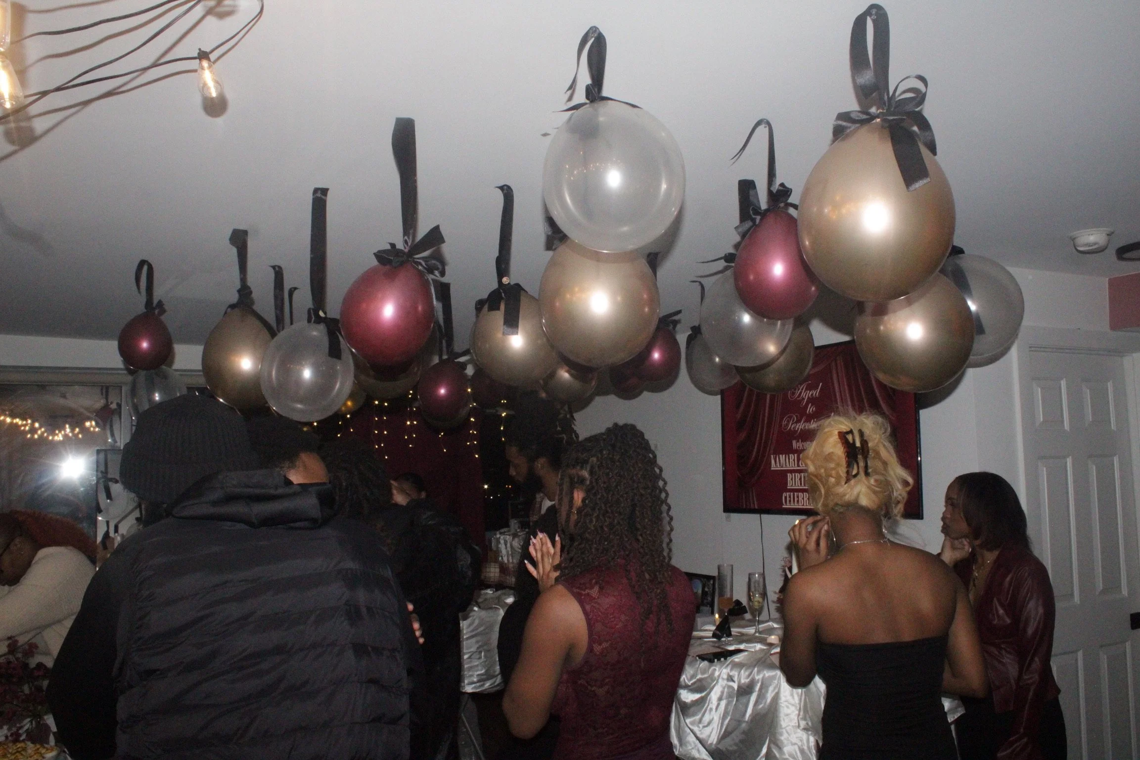 People at a birthday celebration with pink, gold, and clear balloons hanging from the ceiling.