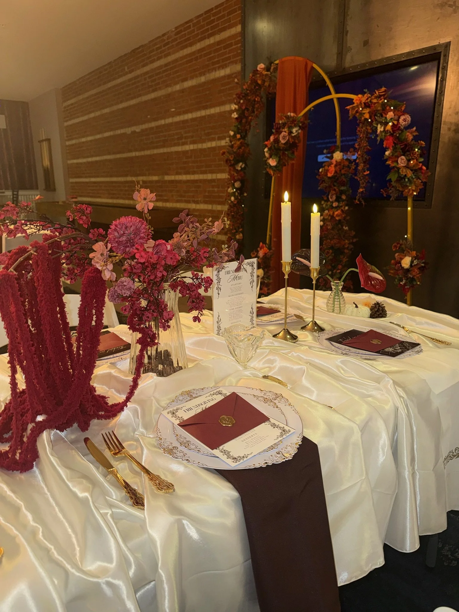 A decorated banquet table with pink and red floral arrangements, white candles, gold cutlery, and a detailed menu. The table is covered with a white satin tablecloth and set for a formal event. Background features a brick wall and a floral arch with 