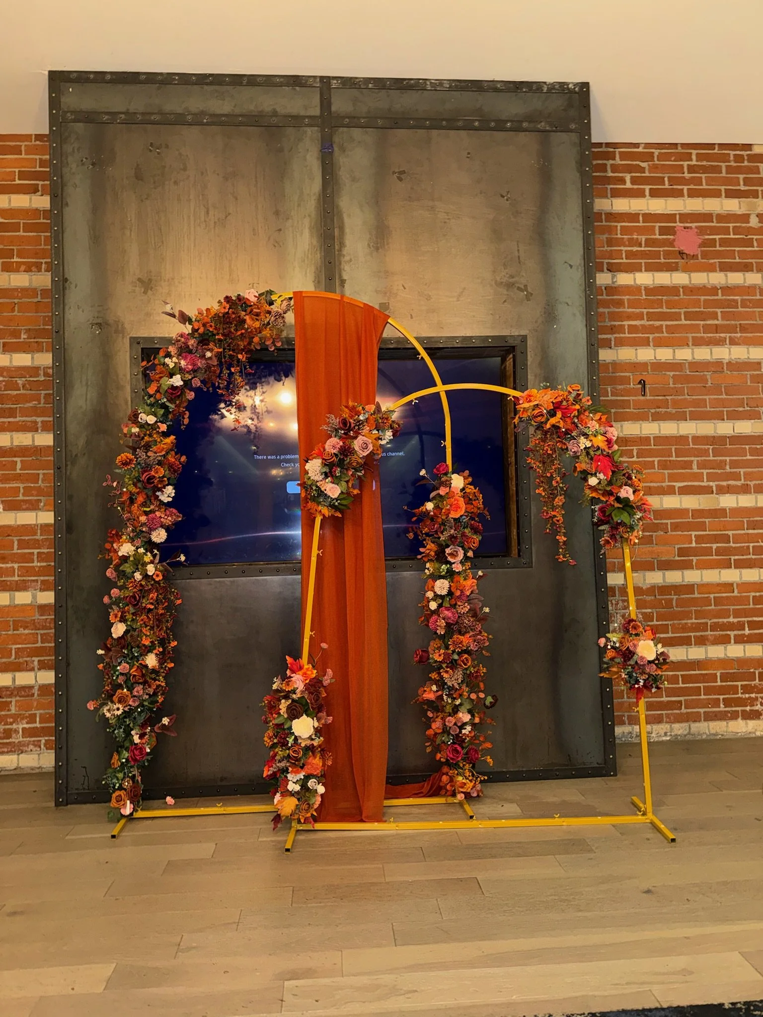 Decorative floral arch with orange fabric and flowers in red, pink, white, and purple, set against a brick wall and gray industrial-style background.