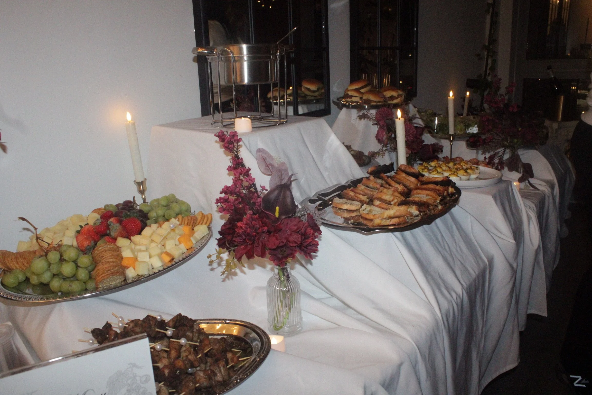 A buffet table with various appetizers, fruits, and snacks, decorated with candles and floral arrangements, set against a white tablecloth and a dimly lit background.