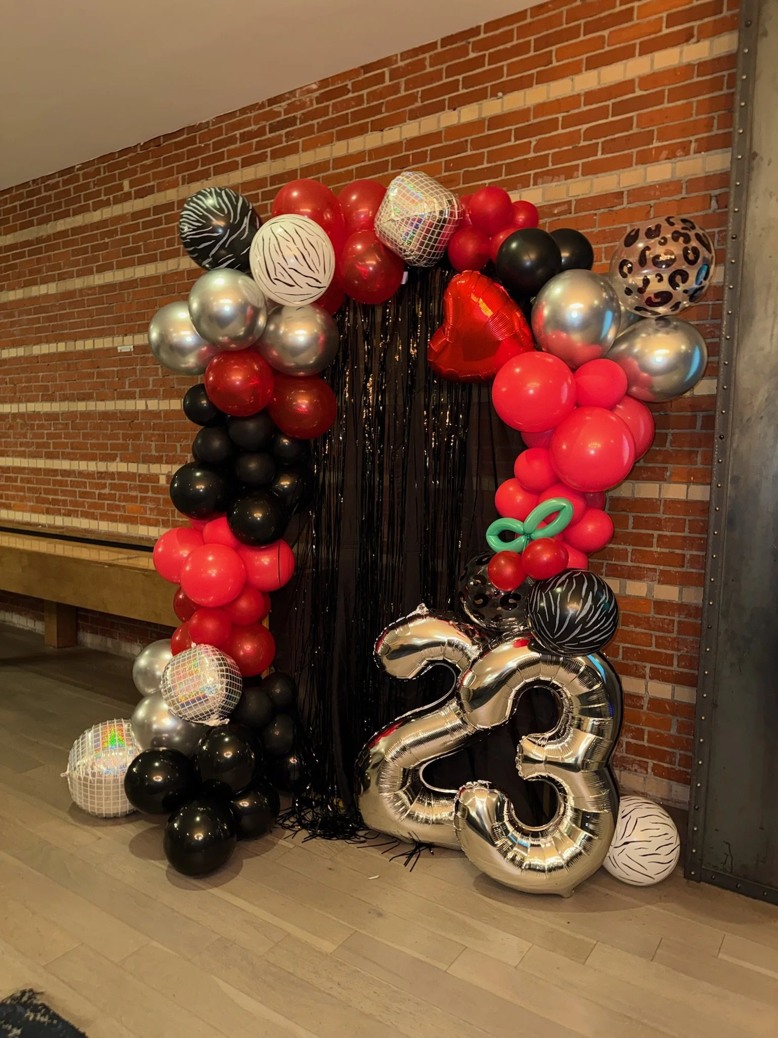 A colorful balloon arch celebrating an 23rd birthday, featuring red, black, silver, and animal print balloons, with large silver '23' numbers and a black backdrop with hanging black fringe.