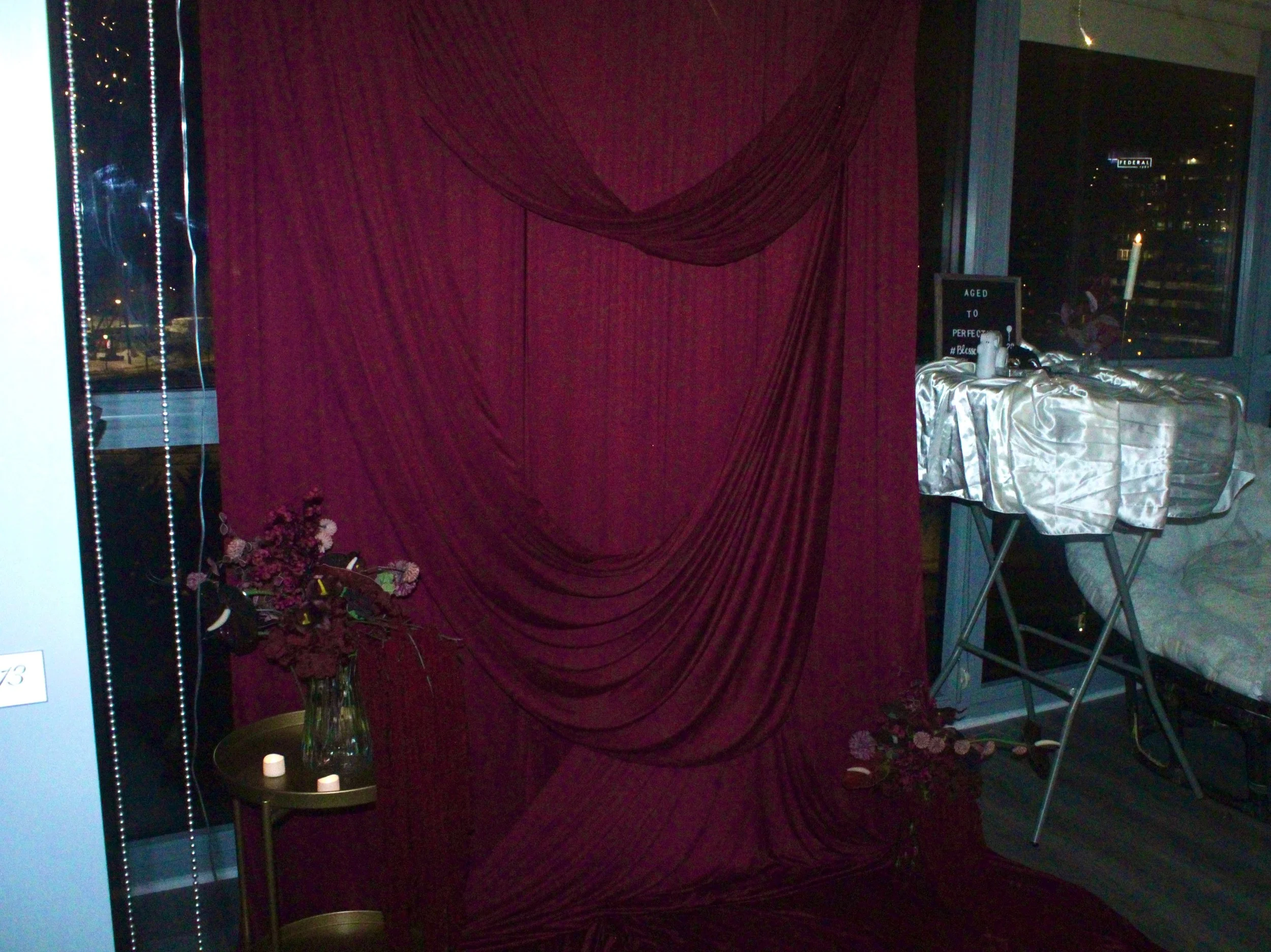 Indoor scene with a burgundy draped fabric backdrop, a small table with flowers and candles, and a table with a satin cloth and a sign indicating the event is aged to perfection, set in a room with large windows showing city lights at night.