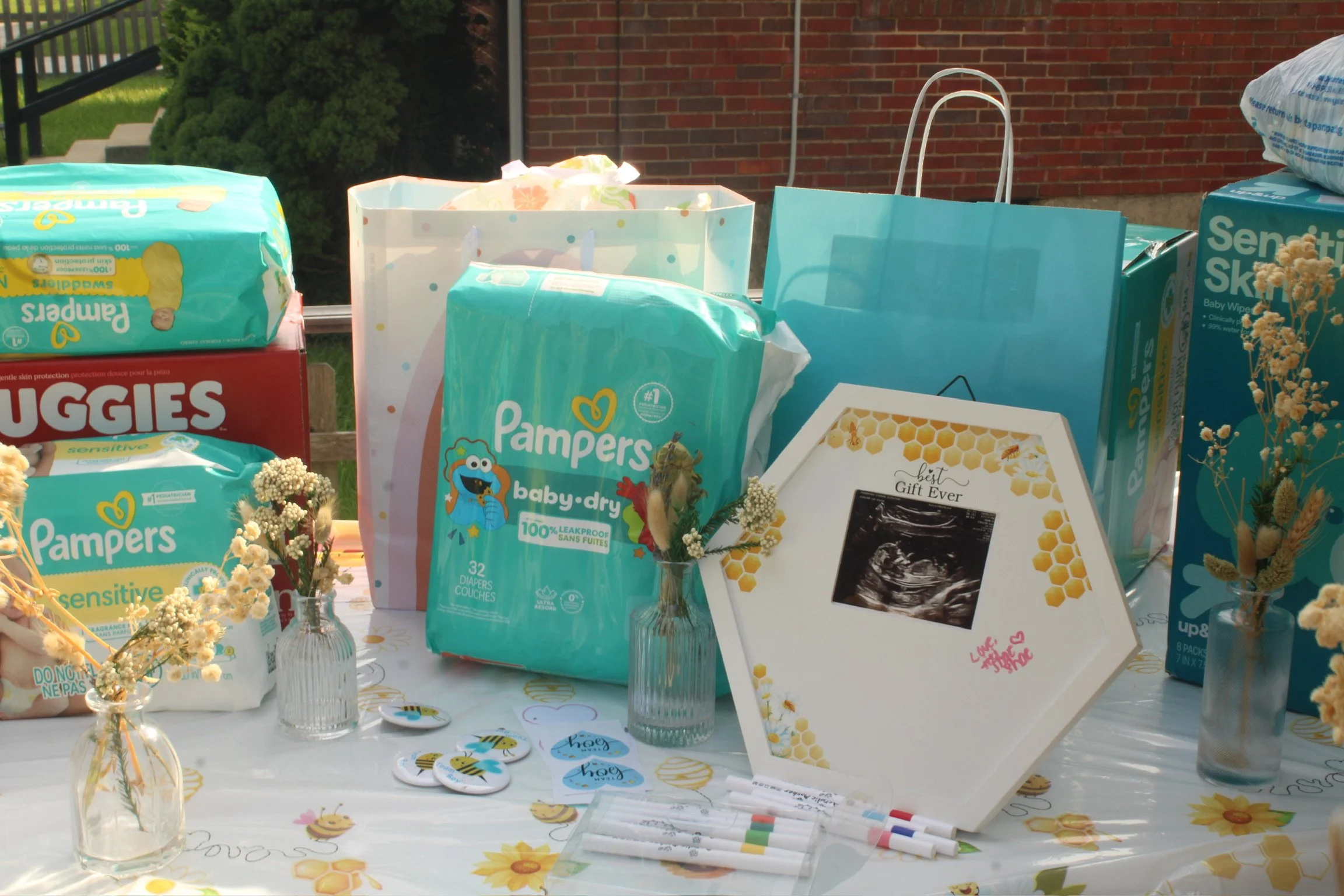 Baby shower table with Pampers diapers, a framed ultrasound photo labeled 'Best Gift Ever', flower vases with baby's breath, and various decorations with a bee theme.