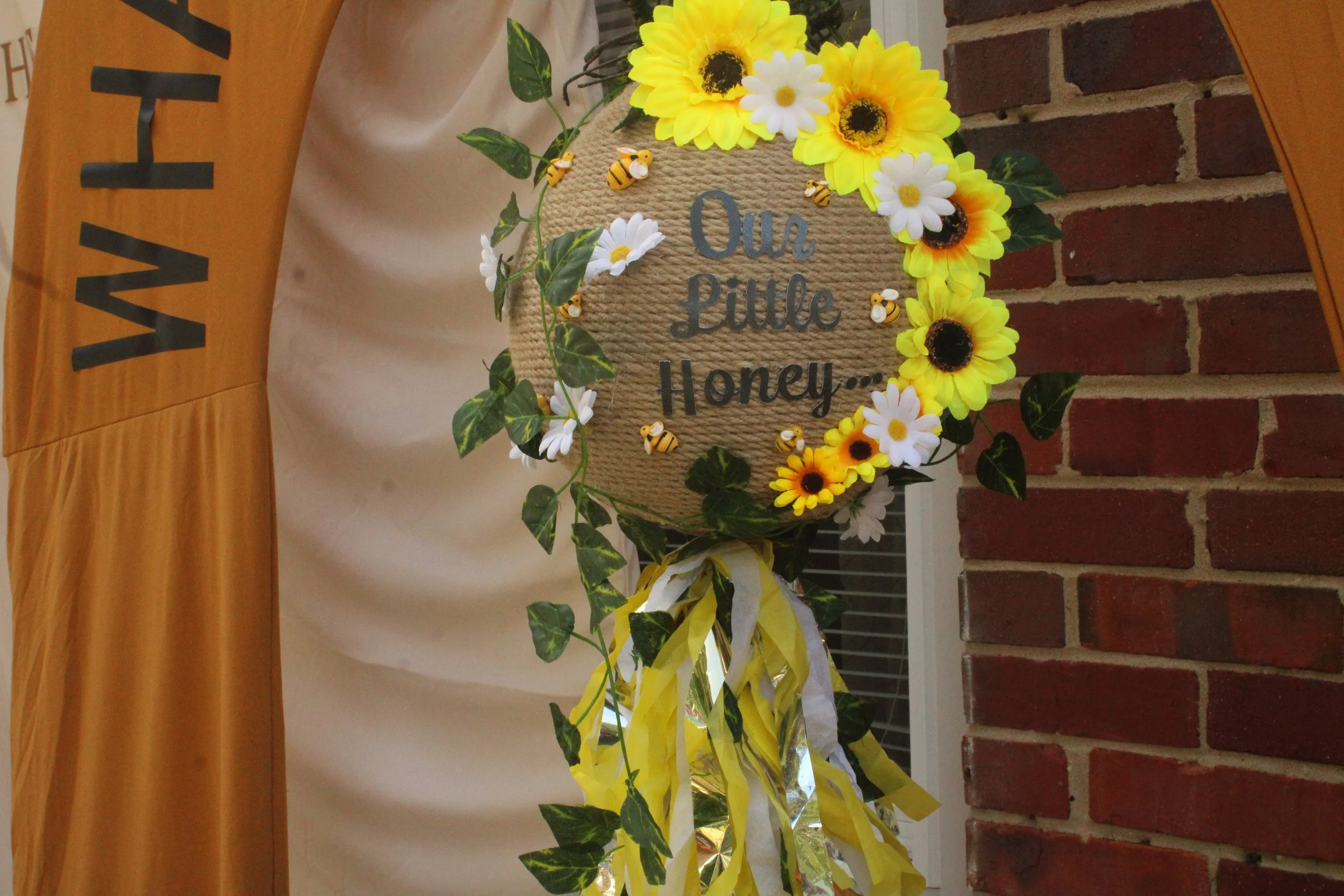 A decorative sign with the words "Our Little Honey..." surrounded by yellow sunflowers, white daisies, small bee embellishments, greenery, and yellow and white ribbons, attached to a brick wall.