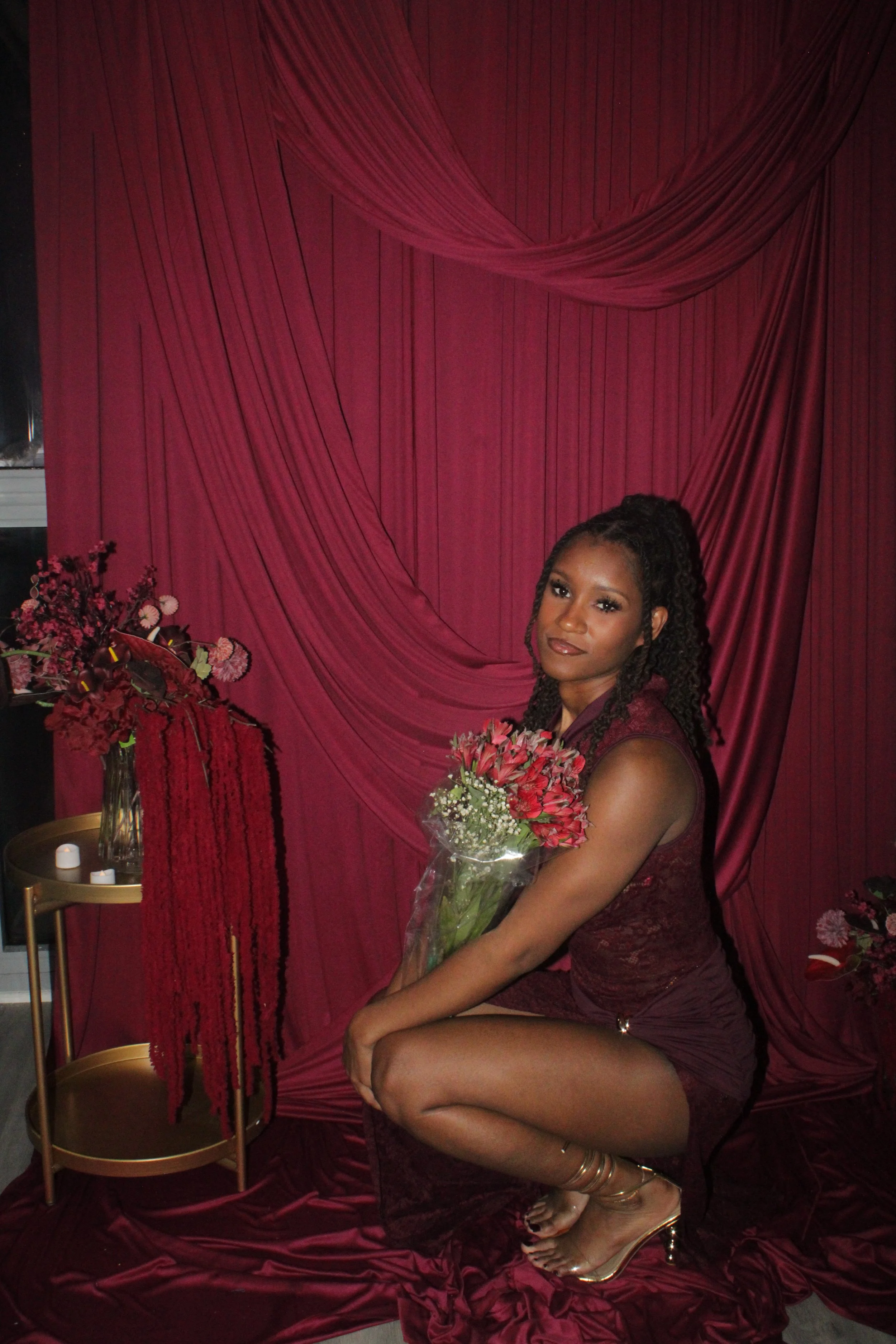 Woman in burgundy dress holding a bouquet of pink flowers, seated against a red draped backdrop with floral arrangements and candles.