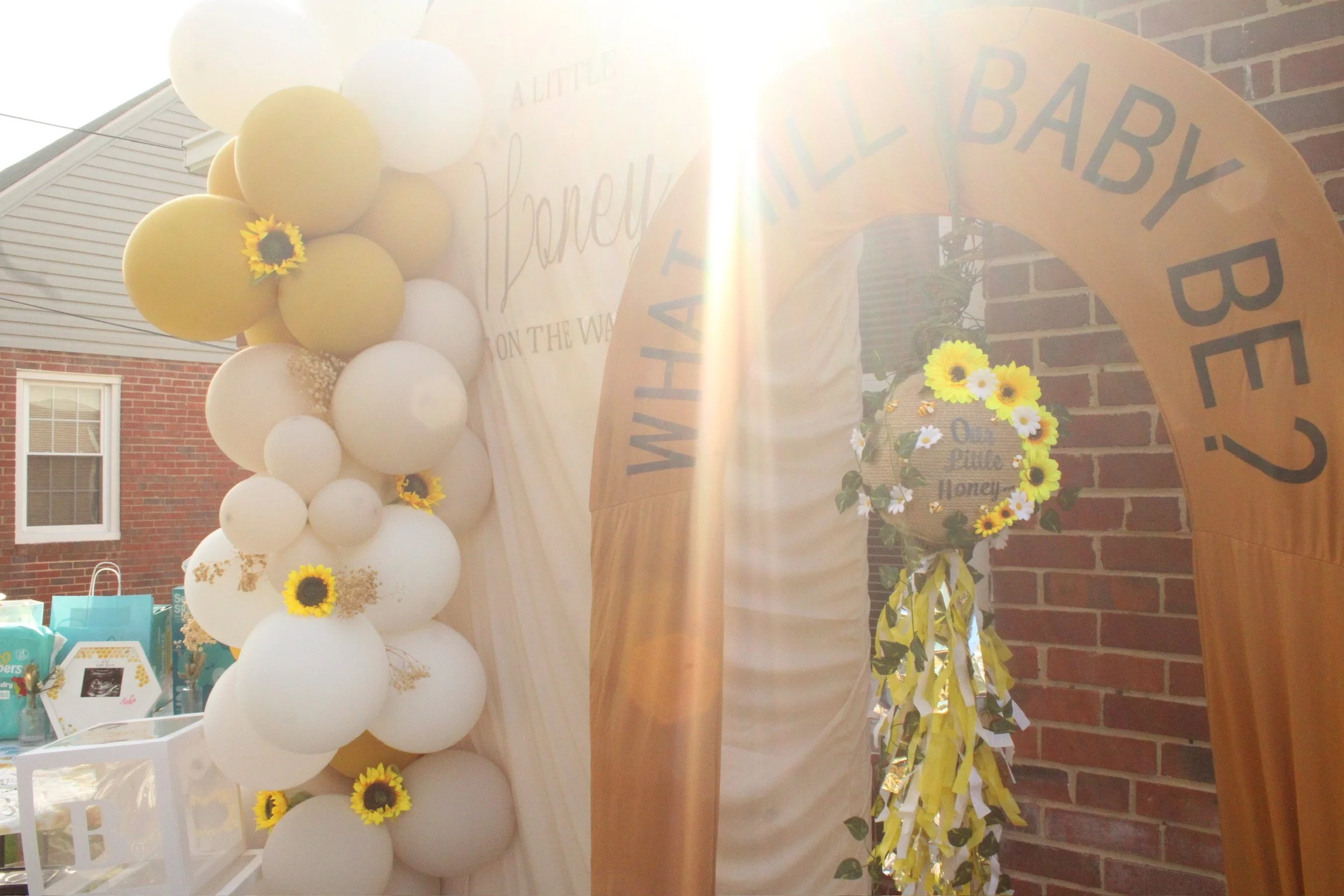 Decorative floral display with white and yellow balloons and sunflowers, set against a brick wall with an arch that says "WANNA BABY?" and a sign that reads "Hello Little Honey".