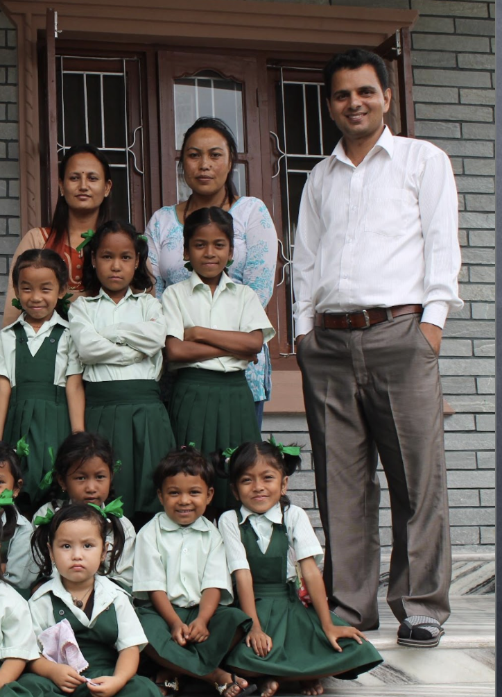 Group of young girls in green school uniforms with green bows, three adult women, and a man standing in front of a brick house with a wooden window frame.