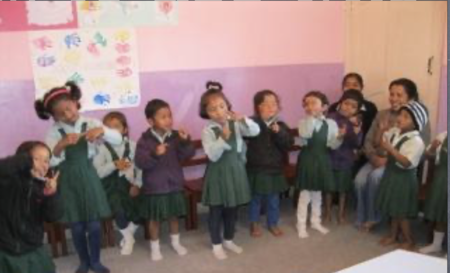 Group of young children in school uniforms, some eating snacks, in classroom with pink and purple walls and colorful artwork.