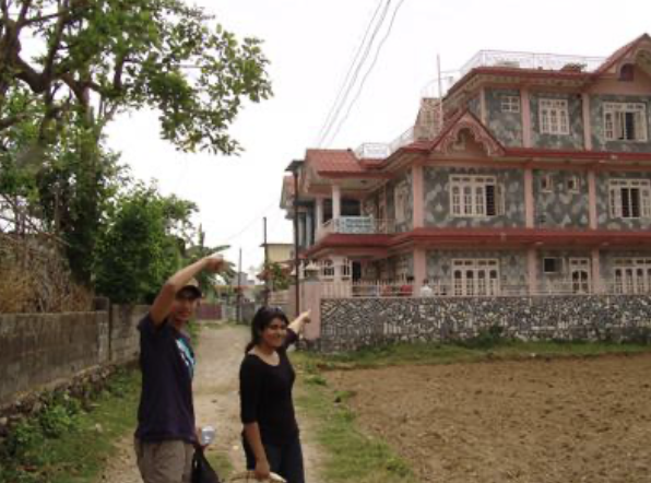 Two women standing on a dirt path in front of a large, ornate residential building with decorative windows and balconies, surrounded by greenery.